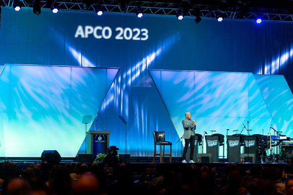 man-standing-on-stage-talking-about-next-generation-9-1-1-upgrades-and-stage-is-lit-up-blue-with-apco-2023-title-at-the-top