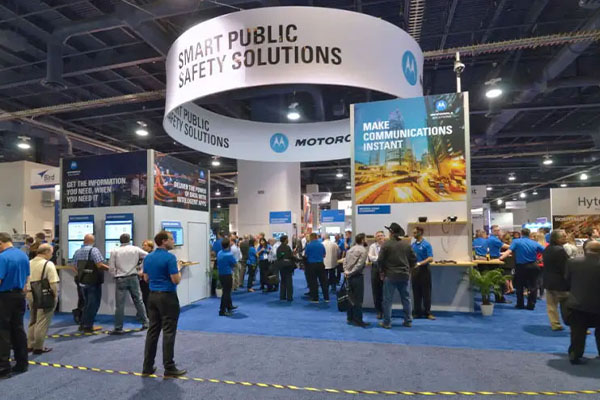 confernce-hall-exhibit-with-giant-hanging-sign-that-says-smart-public-safety-solutions-and-crowd-of-people-below-in-event-booth-talking-about-next-generation-9-1-1-upgrades
