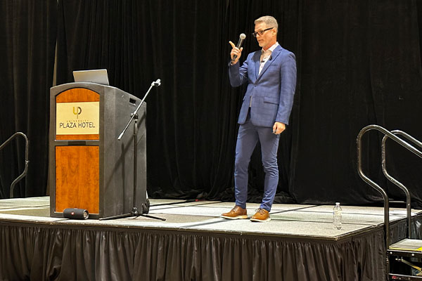 man-in-blue-suit-on-platformed-stage-with-podium-at-missouri-public-safety-communications-conference-in-2023-talking-into-microphone-about-next-generation-911-technology