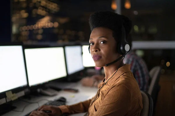 african-american-female-in-tan-shirt-looking-at-camera-with-headset-on-at-a-computer-answering-emergency-next-gen-9-1-1-calls
