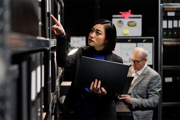 female-with-black-hair-pointing-at-binders-with-laptop-in-hand-to-investigate-next-gen-911-core-services