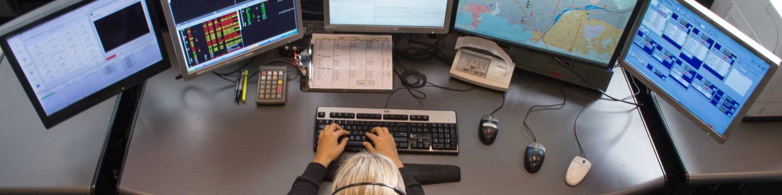 call-taker-sitting-at-desk-in-front-of-multiple-monitors-taking-emergency-calls-and-routing-first-responders-using-next-gen-911-technology