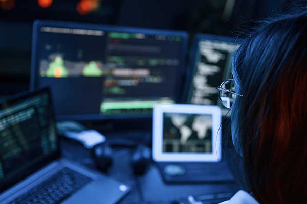 back-of-womans-head-at-her-desk-with-multiple-computer-monitors-in-front-of-her-with-various-graphs-and-code-resembling-next-gen-9-1-1-technology-and-cybersecurity